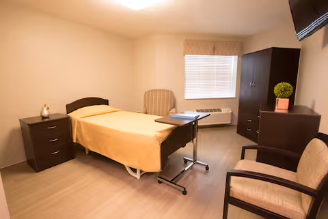 A simple, clean bedroom in a senior living facility with a single bed covered in a yellow blanket, a bedside table with a decorative item, a striped armchair, a small overbed table, a dark wooden dresser with a potted plant, a wall-mounted TV, and a window with blinds and a valance.