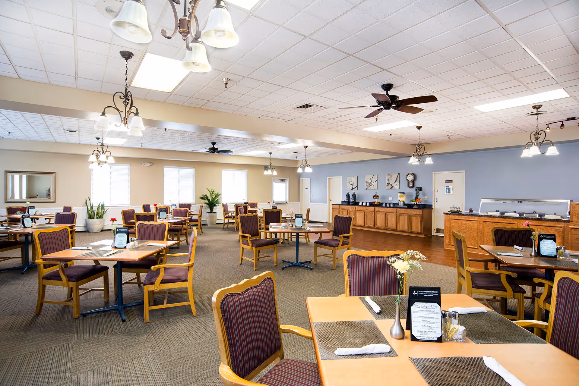 A spacious dining room with multiple wooden tables and chairs with striped upholstery. The tables are set with placemats, napkins, menus, and small flower vases. The room has a carpeted floor, ceiling fans, and hanging light fixtures. There is a buffet area with wooden cabinets and beverage dispensers along one wall, and large windows letting in natural light.