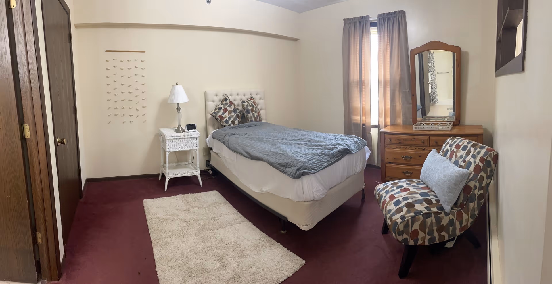 A small bedroom with a single bed covered with a gray quilt and two patterned pillows. Next to the bed is a white wicker nightstand with a lamp and a digital clock. There is a wooden dresser with a mirror and a patterned upholstered chair with a white decorative pillow. The room has beige walls, a window with beige curtains, and a maroon carpet with a small white rug.