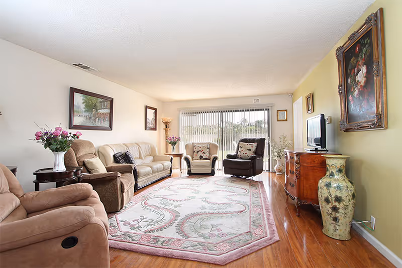 A bright and spacious living room with wooden flooring, a large decorative area rug, multiple upholstered armchairs and a sofa, a wooden dresser with a flat-screen TV, floral paintings on the walls, and large sliding glass doors with vertical blinds letting in natural light.