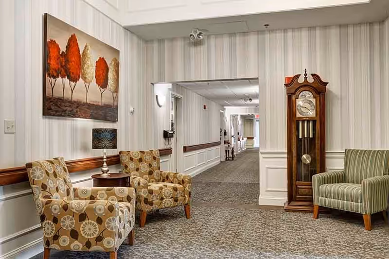 A hallway in a senior living facility with patterned carpet and striped wallpaper. There are two patterned armchairs and a small round wooden table with a lamp on it on the left side. On the right side, there is a tall wooden grandfather clock and a green striped armchair. A painting of trees with red and white foliage hangs on the left wall above the chairs. The hallway extends into the distance with doors and light fixtures visible.