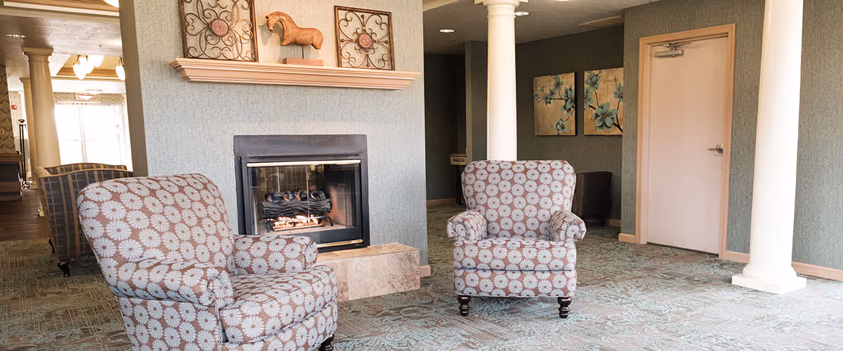 A cozy seating area in a senior living facility featuring two patterned armchairs facing a fireplace with decorative items on the mantel. The room has carpeted flooring, columns, and artwork on the walls, creating a comfortable and inviting atmosphere.