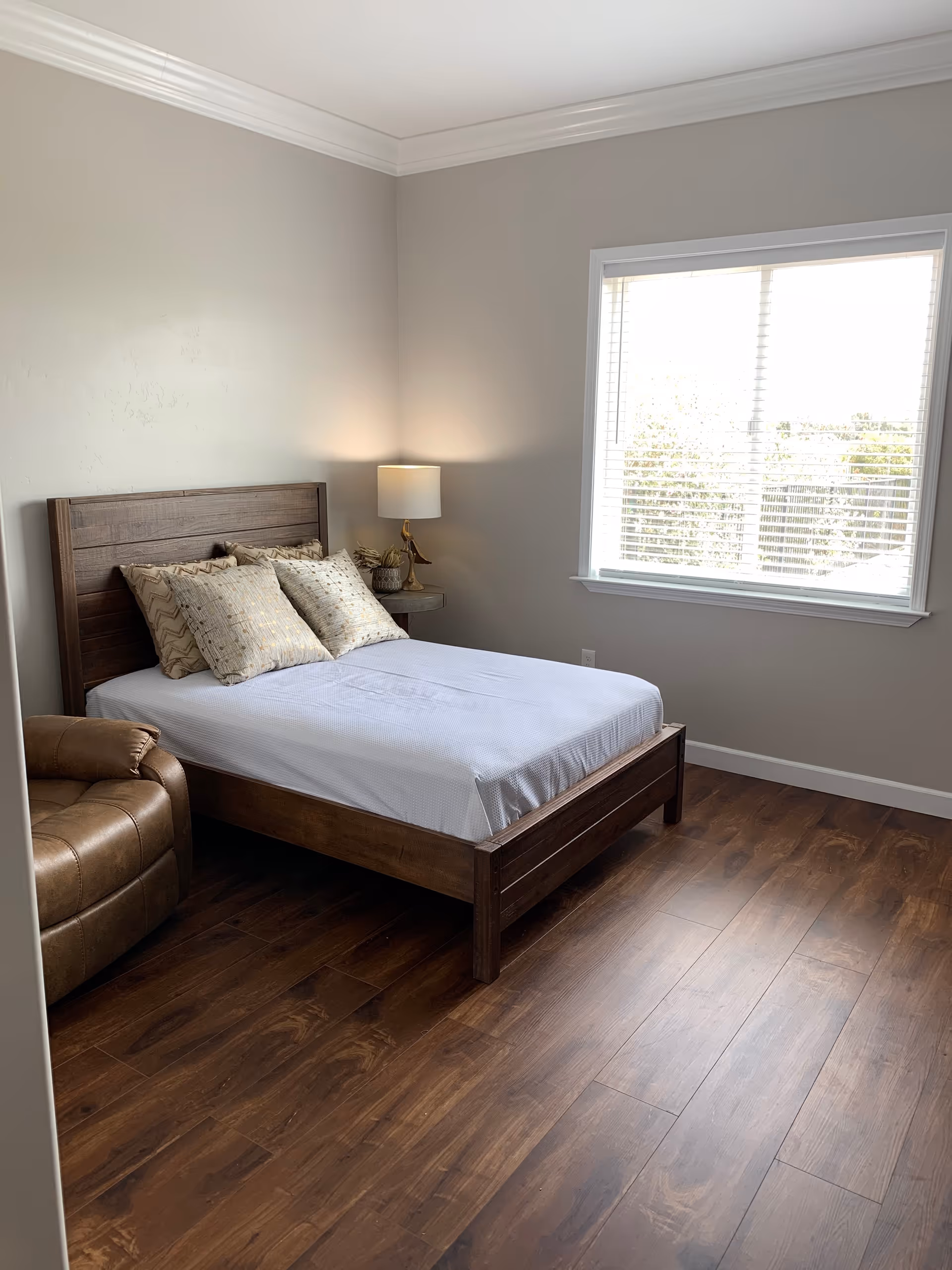 A bright bedroom with a wooden bed topped with pillows, a bedside table and lamp, a leather chair, and a window with blinds.