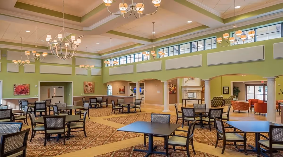 A spacious dining area with multiple tables and chairs arranged on a patterned carpet. The room has high ceilings with several chandeliers and large windows near the ceiling allowing natural light. The walls are painted green with white trim, and there are decorative paintings on the walls. In the background, there is a seating area with armchairs and a fireplace.