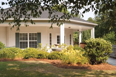 Exterior view of a single-story building with white siding and green shutters, surrounded by well-maintained bushes, plants, and a small lawn. A covered porch with white columns is visible, along with a concrete walkway and trees in the background.