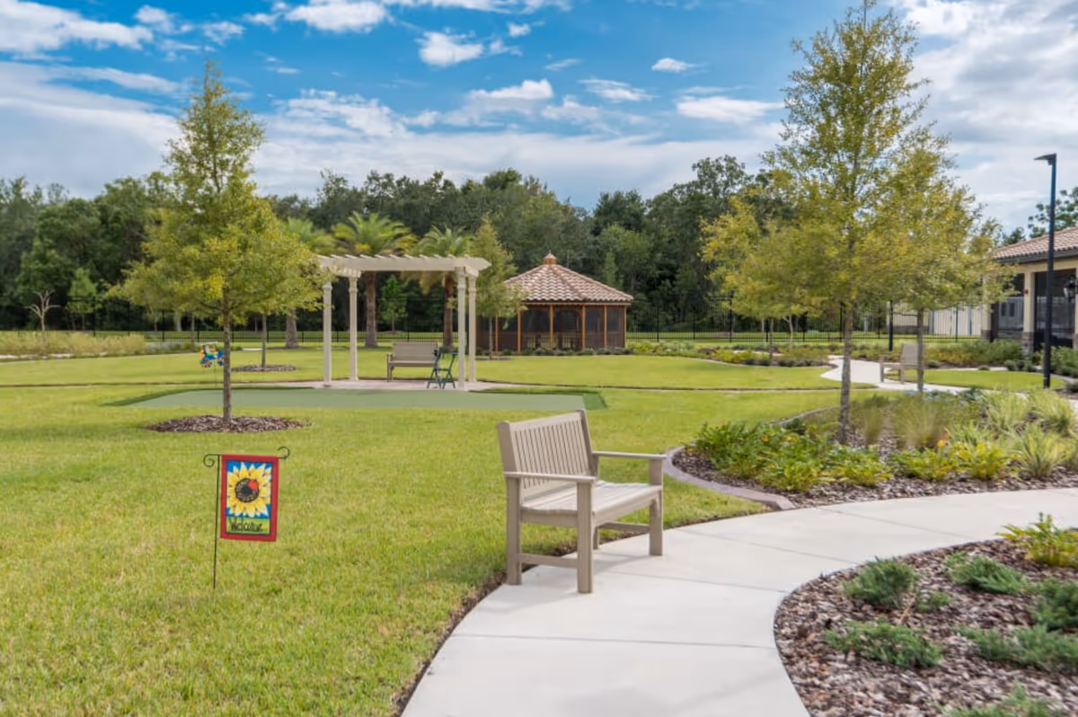 A well-maintained outdoor garden area with green grass, trees, and a curved concrete pathway. There are several benches along the path, a small pergola with a bench underneath, and a gazebo with a tiled roof in the background. A colorful welcome sign featuring a sunflower is placed in the grass near the pathway. The sky is partly cloudy with blue patches visible.
