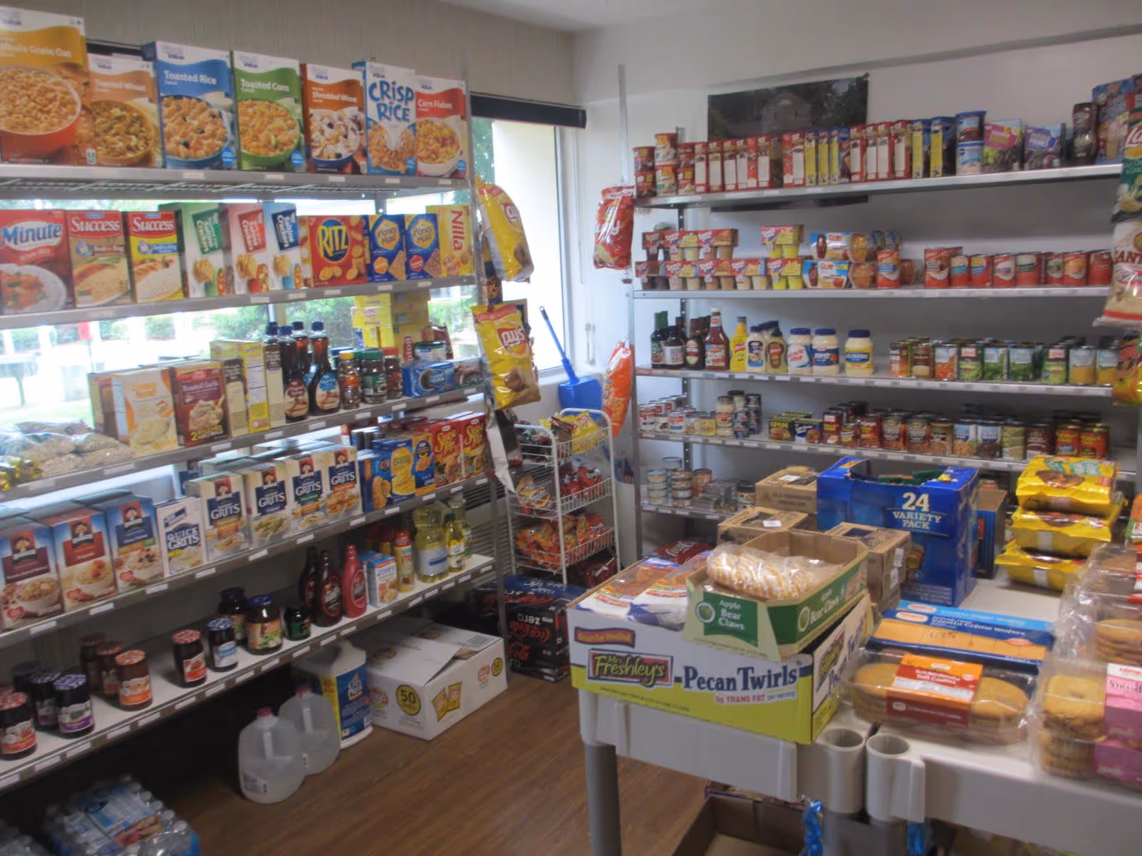 Interior view of a small grocery or pantry area with shelves stocked with various food items including cereals, canned goods, condiments, snacks, and bottled water. There is a table in the center with boxes of packaged food items. A window is visible in the background letting in natural light.
