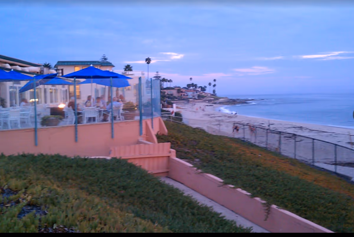 Outdoor dining patio with blue umbrellas and people sitting at tables overlooking a sandy beach and ocean at dusk.