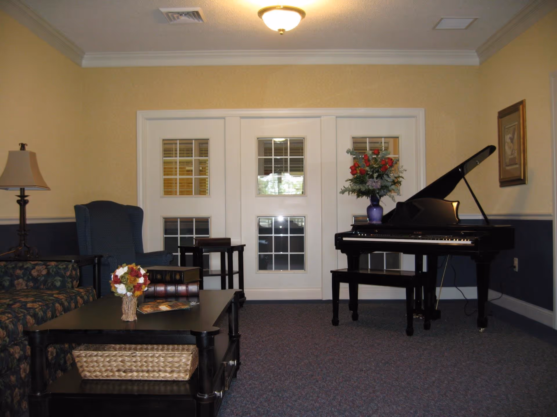 A cozy living room area with a floral patterned sofa, a blue armchair, a black coffee table with a small flower arrangement and books, a floor lamp, and a black grand piano with a vase of red and white flowers on top. The room has beige and blue walls, a carpeted floor, and white double doors with glass panes.