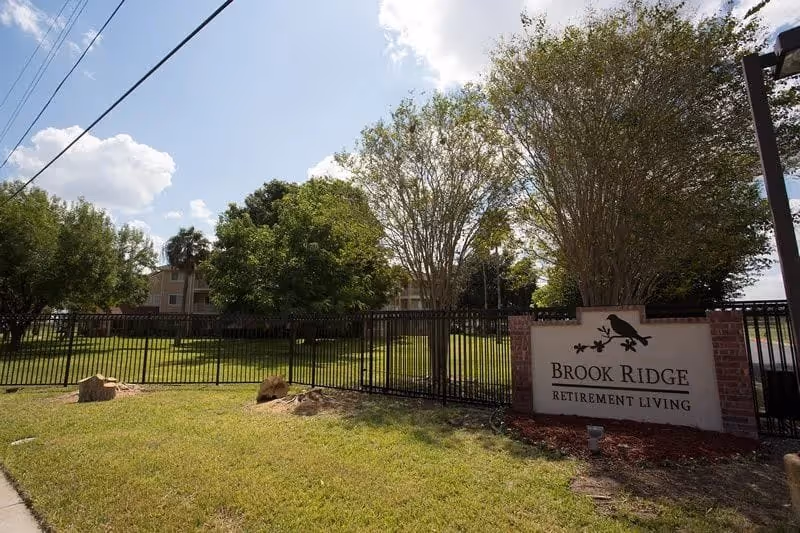 Outdoor view of Brook Ridge Retirement Living facility showing a grassy area with trees, a black metal fence, and a sign with the facility's name. The sky is partly cloudy.