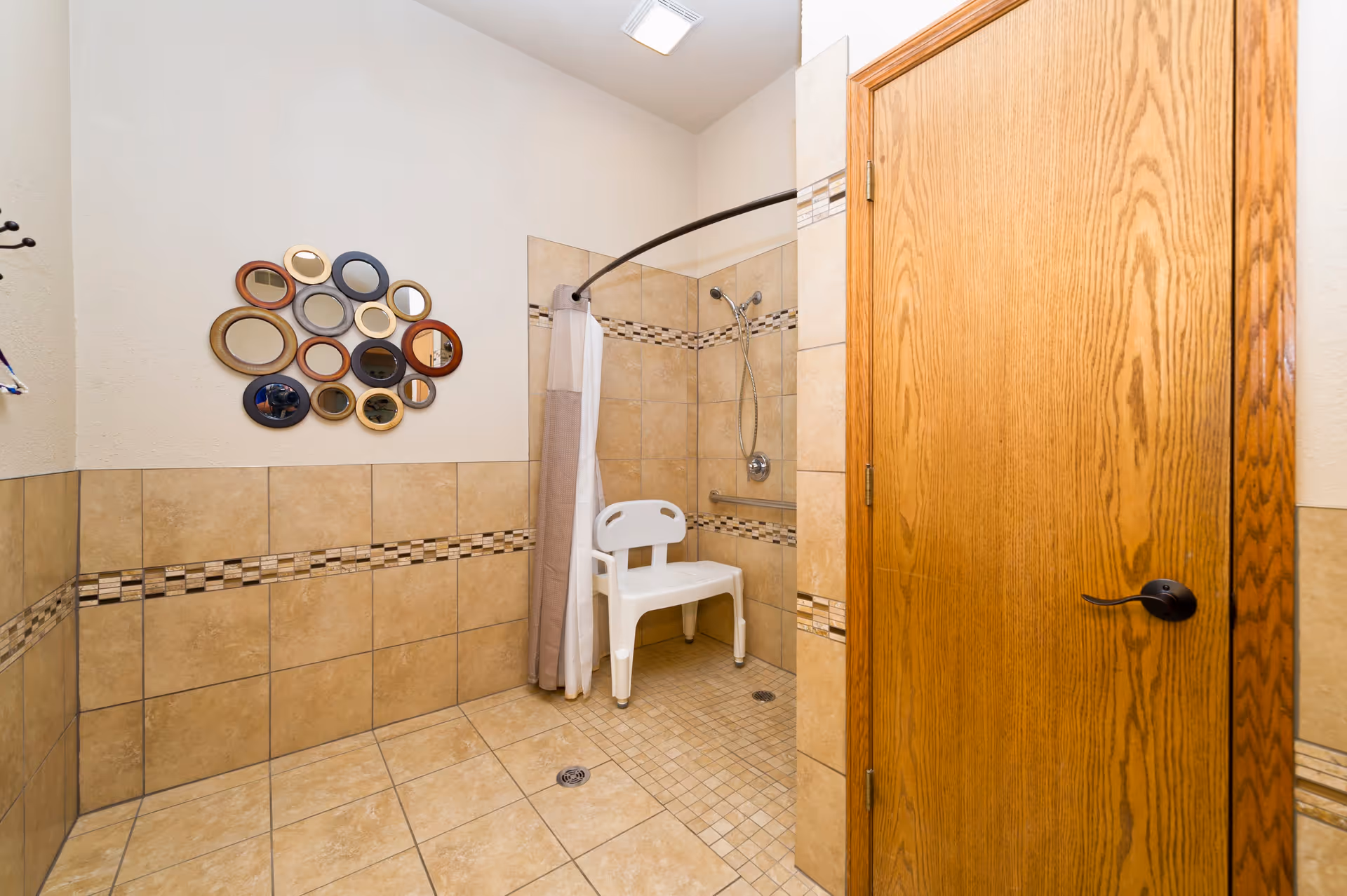 A tiled bathroom with a walk-in shower featuring a white plastic shower chair and a curved shower curtain rod with a beige and white shower curtain. The walls have beige tiles with a decorative mosaic tile border. A wooden door with a black handle is partially visible on the right. On the left wall, there is a decorative arrangement of multiple small round mirrors with different colored frames.