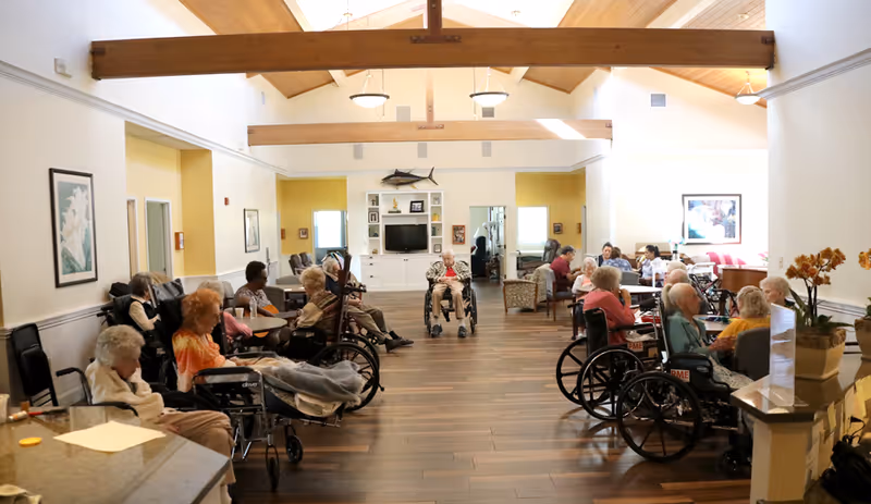 A spacious common area in an assisted living community with wooden beams on the ceiling and hardwood floors. Several elderly residents, many in wheelchairs, are seated around tables and in chairs, engaging in conversation or resting. The room is well-lit with natural light coming through windows. There is a TV mounted on a white cabinet against the far wall, and framed artwork on the walls.