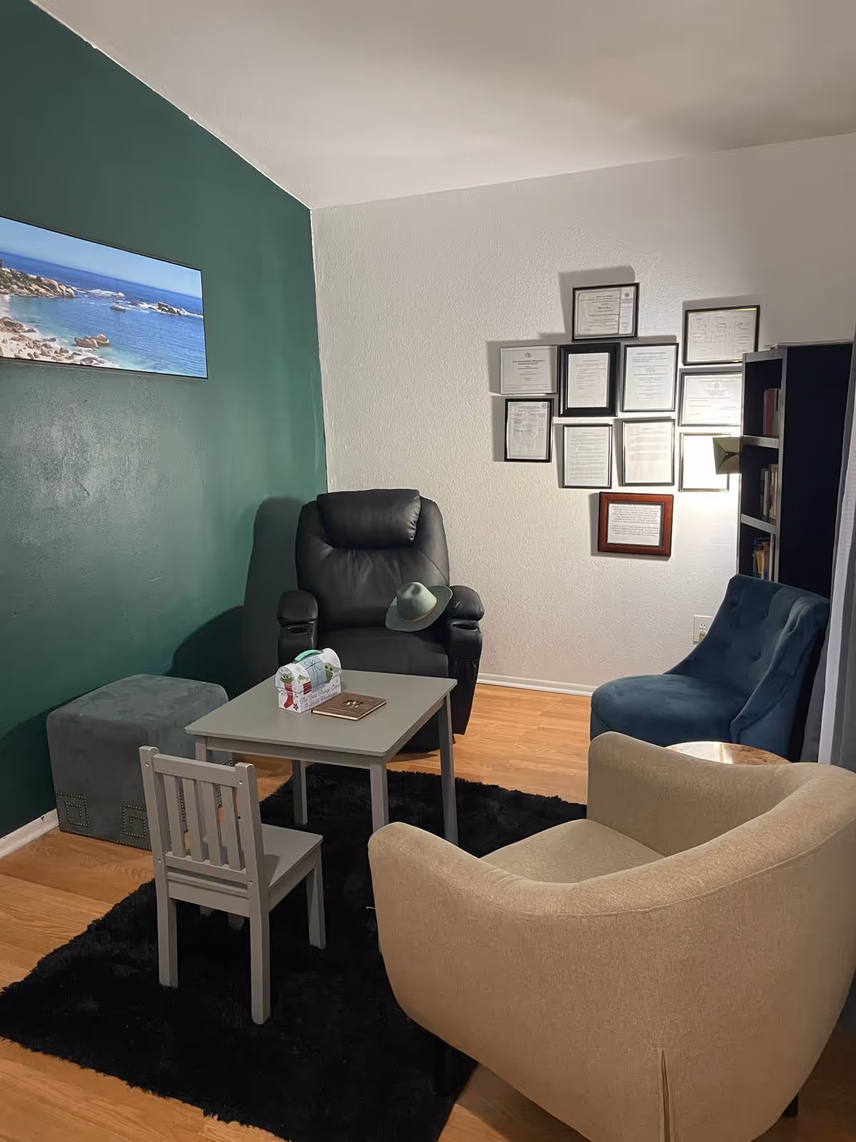 A cozy sitting area with a black leather recliner, a beige armchair, a blue upholstered chair, a small gray table with a tissue box and a book, and a small gray chair. The walls are painted green and white, with framed certificates or documents on the white wall and a landscape picture on the green wall. The floor is wooden with a black rug underneath the furniture.