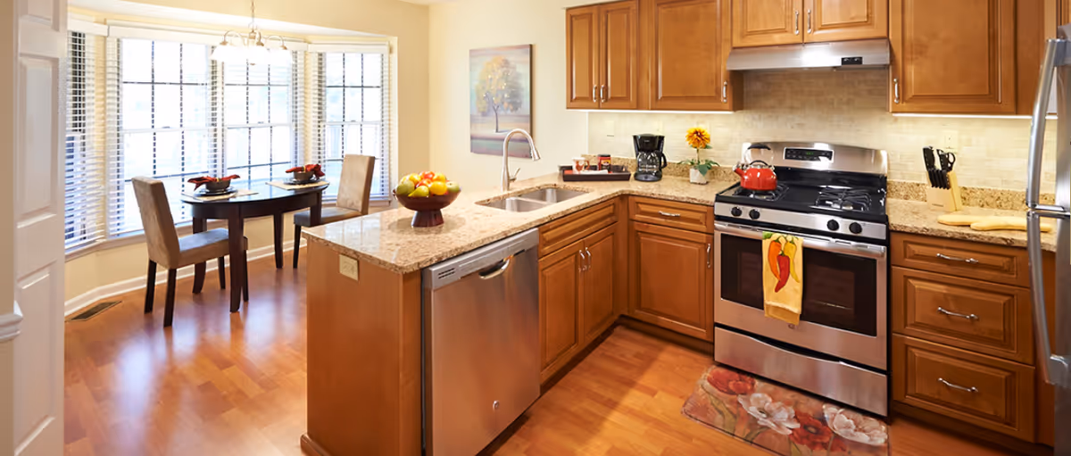 A bright kitchen with wooden cabinets, granite countertops, and stainless steel appliances including a dishwasher, stove, and refrigerator. A small dining area with a table set for two is visible near large windows with blinds. The kitchen has a wooden floor and a colorful rug in front of the stove.