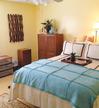 A cozy bedroom with a neatly made bed featuring white and brown pillows and a light blue blanket. There is a wooden cabinet with decorative items on top, a small chest of drawers, a wall-mounted wooden art piece, and a floor lamp providing warm light.