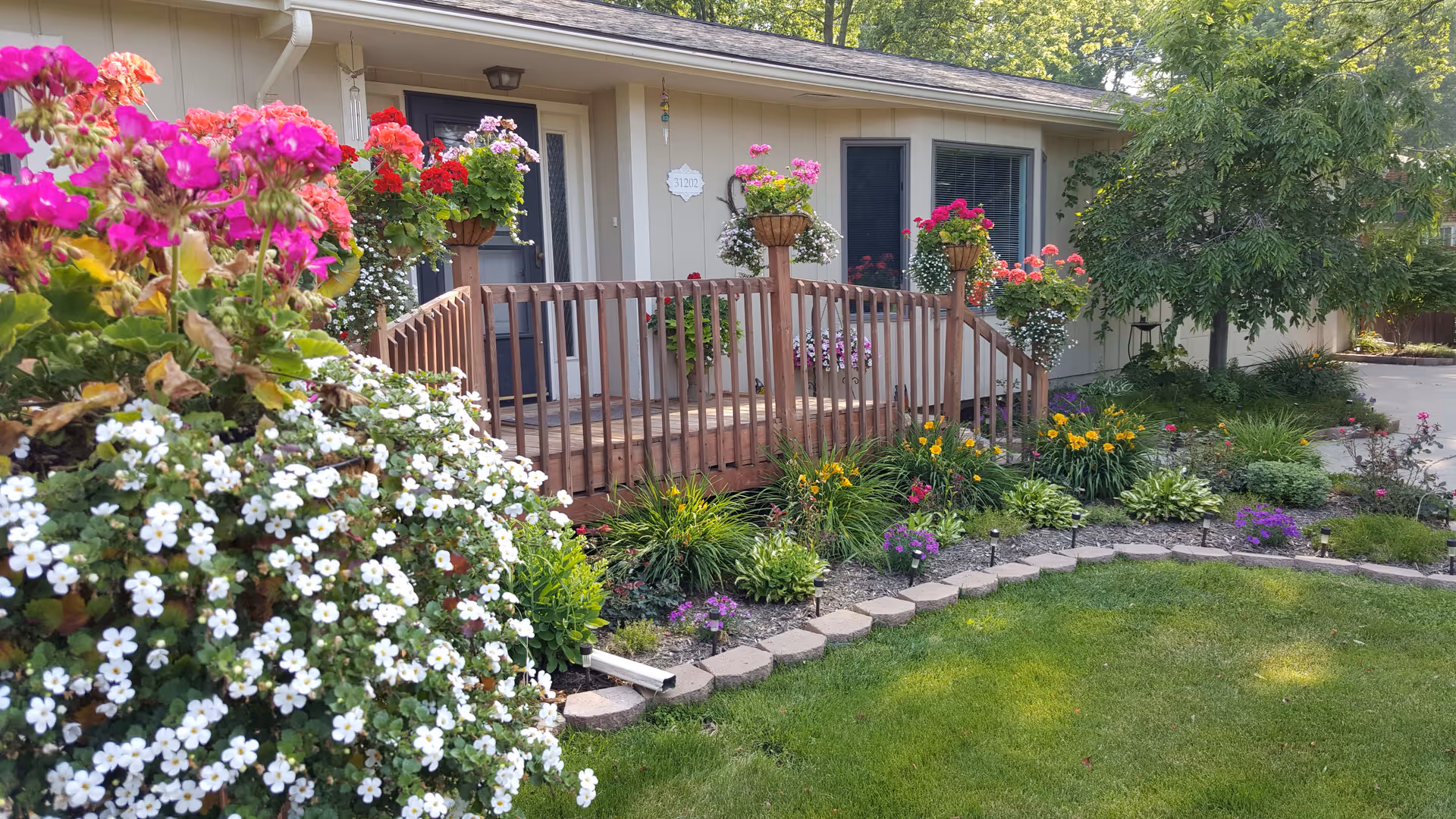 A well-maintained garden area in front of a single-story building with a wooden ramp leading to a door. The garden features vibrant flowers in various colors including pink, red, white, yellow, and purple, along with green shrubs and a neatly trimmed lawn. Trees and additional greenery surround the area, creating a peaceful outdoor environment.