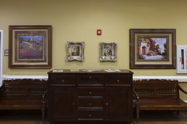 Interior wall with four framed paintings hung above a dark wooden cabinet. Two wooden benches are placed on either side of the cabinet. The wall is painted light yellow and has a fire extinguisher cabinet on the right side.