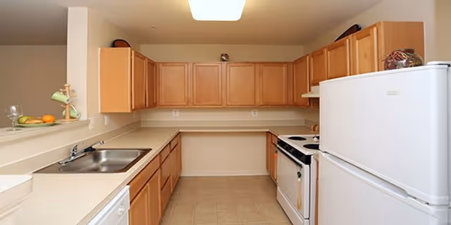 A clean kitchen with light wood cabinets, beige countertops, a white refrigerator, a white stove with oven, a stainless steel sink, and tiled floor. There are some fruits and a decorative item on the counter.