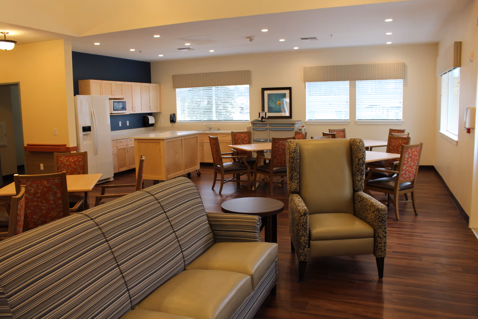 Open communal living and dining area with sofas and an armchair in the foreground, dining tables and chairs, and a kitchenette in the background.