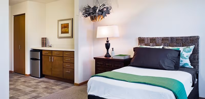 A bedroom in a senior living facility featuring a bed with white and green bedding, a dark wooden nightstand with a lamp and a decorative hanging light fixture above. To the left, there is a small kitchenette area with wooden cabinets, a mini refrigerator, and a closed wooden door.
