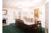 Formal dining room with a long wooden table and chairs, chandelier, fireplace, painting, and green carpet.