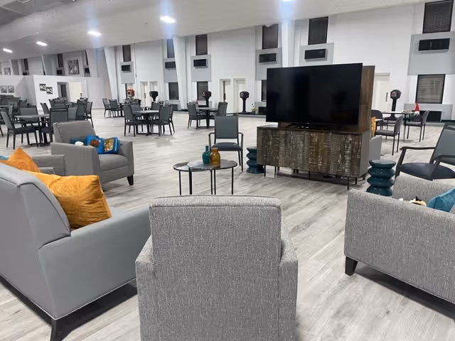 Spacious common area in a senior living facility with multiple seating arrangements including gray armchairs and sofas with orange and blue cushions, small round tables, and a large flat-screen TV on a wooden stand. The room has light wood flooring, white walls, and several windows with blinds.