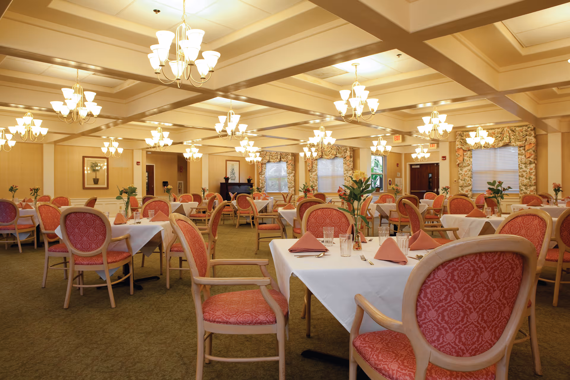 Spacious dining room with multiple tables set with white linens, pink-upholstered chairs, chandeliers, and floral centerpieces.