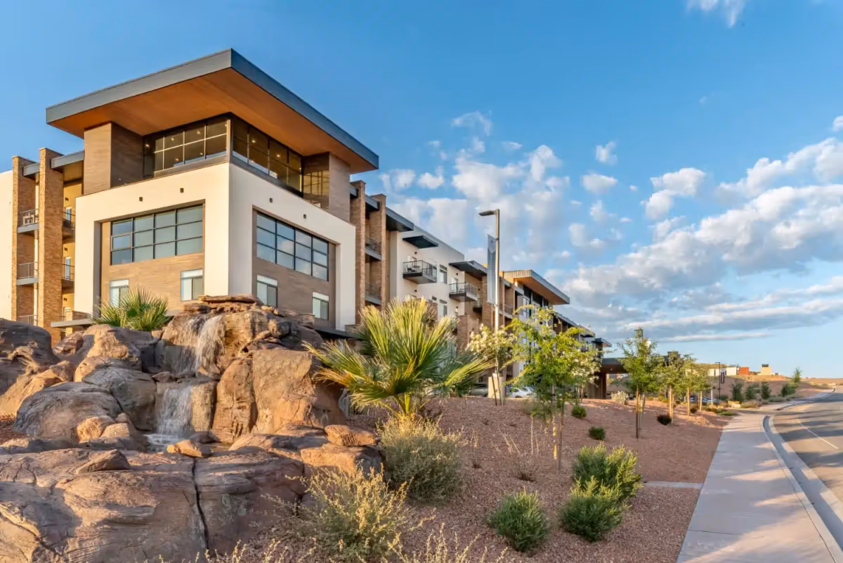 Modern multi-story building with rock waterfall landscaping, desert plants, and a sidewalk under a blue sky.