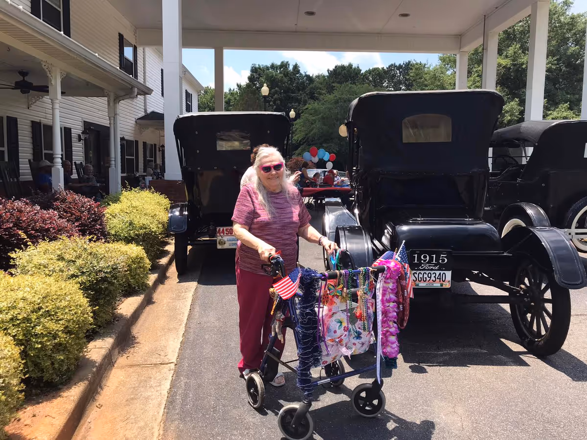 An elderly woman wearing pink sunglasses and a purple outfit stands outside a senior living facility using a decorated walker with American flags and colorful accessories. Behind her are two vintage black cars parked under a covered driveway, with other people sitting and socializing in the background near a table with balloons. The building has white siding and black shutters with bushes lining the walkway.