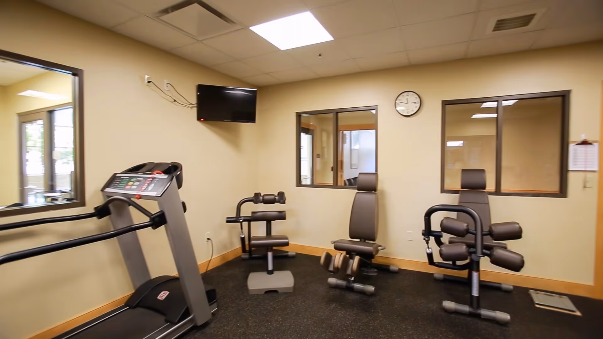 A small fitness room with exercise equipment including a treadmill, a leg curl machine, and a leg extension machine. The room has beige walls, a wall-mounted TV, two windows looking into another room, a clock on the wall, and a rubberized floor.