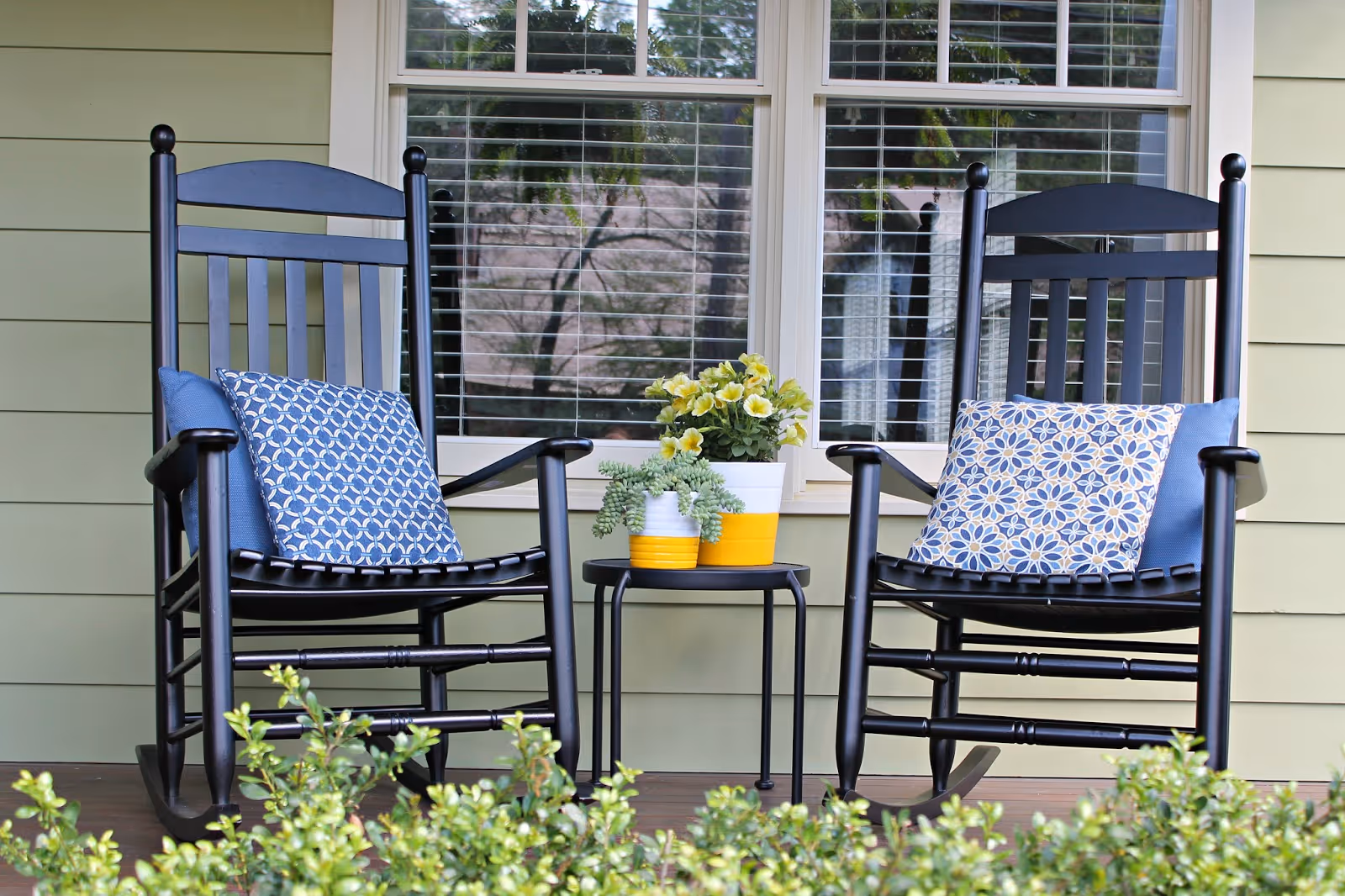 Two black rocking chairs with blue patterned cushions and solid blue pillows on a porch with a small black table between them holding two potted plants, in front of a window with white blinds and green siding walls.
