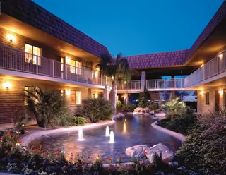 Evening view of a courtyard at Wooddale Village Retirement featuring a central pond with small illuminated fountains, surrounded by lush greenery and palm trees. The two-story building with balconies and warm exterior lighting encloses the courtyard.