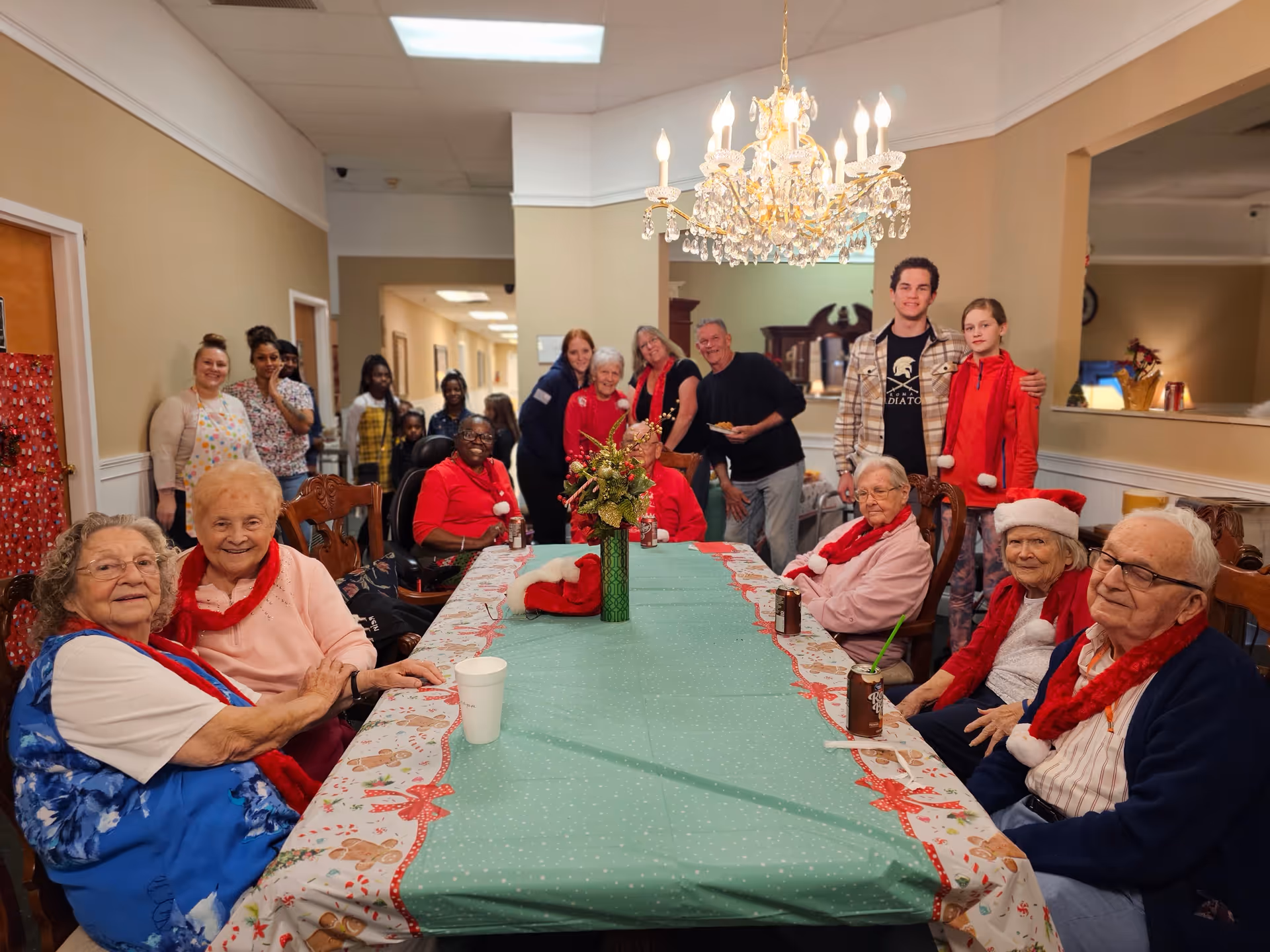 A group of elderly people and some younger visitors gathered around a long table decorated with a festive tablecloth and a centerpiece. The elderly individuals are seated, some wearing red scarves and holiday hats, while the younger visitors stand behind them. The room has beige walls, a chandelier overhead, and a warm, cheerful atmosphere.