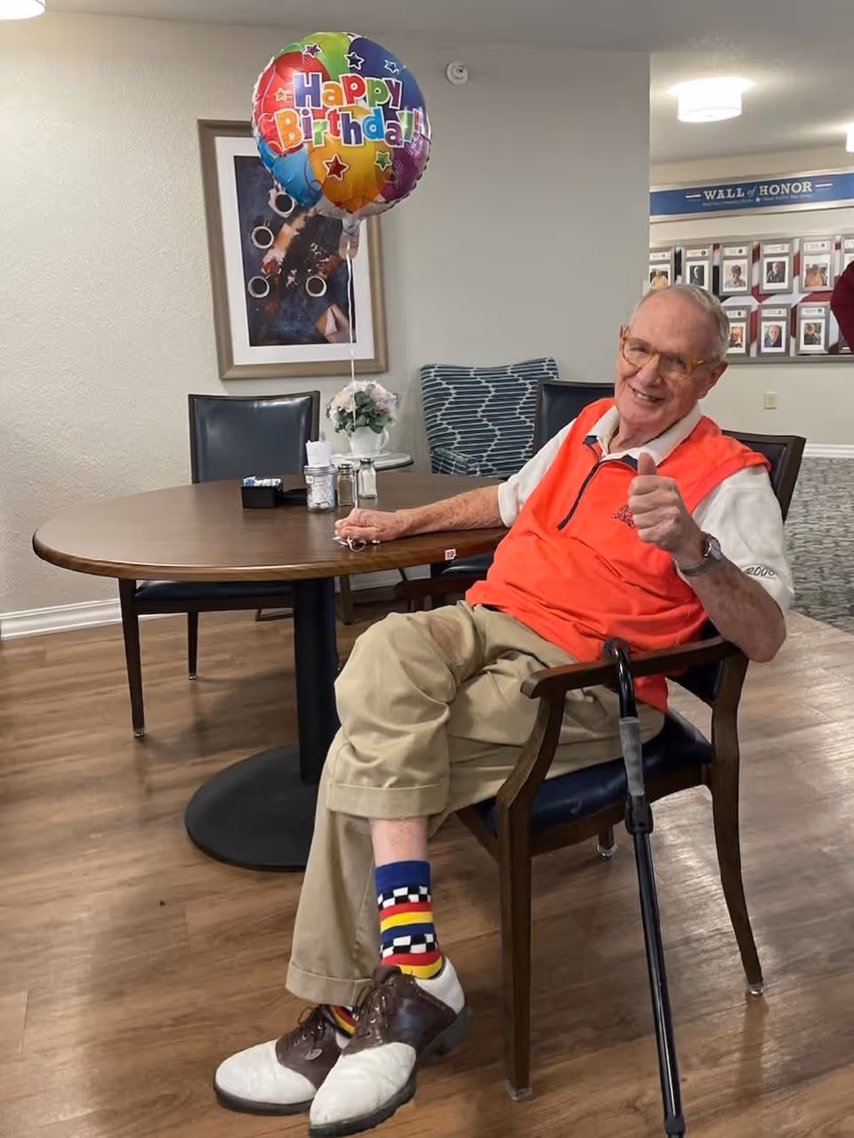 An elderly man sitting on a chair in a senior living facility, giving a thumbs-up. He is wearing a bright orange vest, beige pants, colorful socks, and brown and white shoes. A cane rests against his chair. On the table next to him is a colorful 'Happy Birthday' balloon, a small flower arrangement, and some condiments. The room has wooden flooring, framed artwork on the wall, and a 'Wall of Honor' display in the background.
