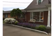 Exterior view of a brick senior living facility with a large window, a white bench in front, and landscaping including bushes and flowering plants along the building's edge.