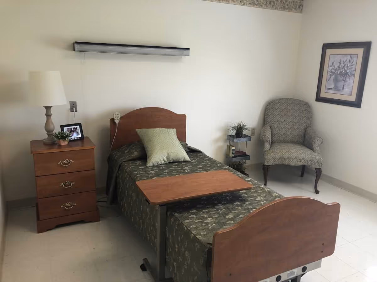 A simple nursing home bedroom with a single bed covered in a green patterned bedspread and a matching pillow. There is a wooden bedside table with three drawers, a lamp, a small plant, and a framed photo. A small rolling overbed table is positioned over the bed. In the corner, there is an upholstered armchair with a patterned fabric and a framed picture hanging on the wall above it. The room has plain white walls and a tiled floor.