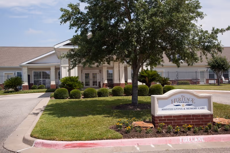 Exterior view of The Heritage Tomball Senior Living facility showing the front entrance with a manicured lawn, bushes, a tree, and a sign that reads 'The Heritage Tomball Assisted Living & Memory Care'.