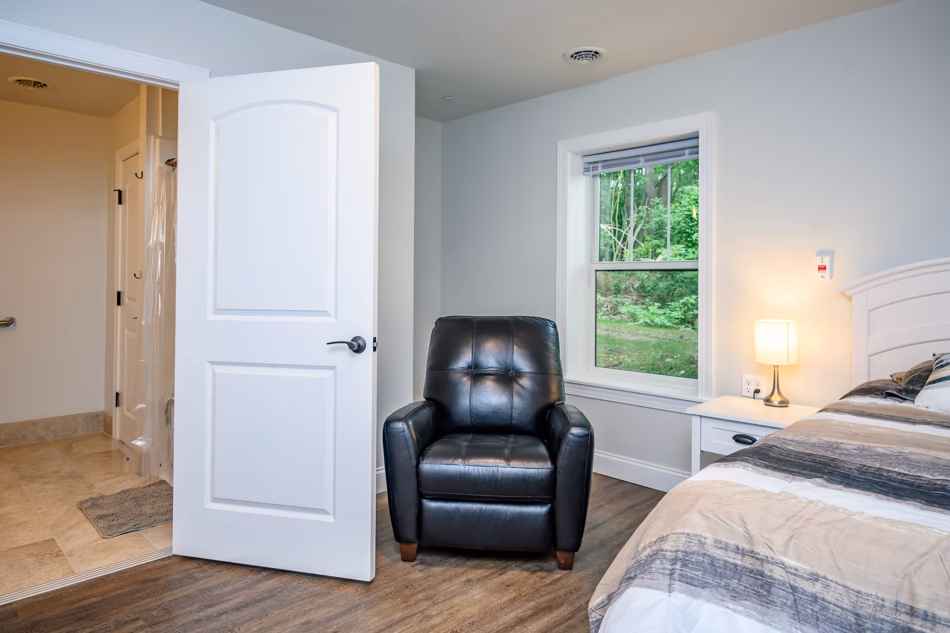 A cozy bedroom in an assisted living facility featuring a black leather armchair, a bed with a striped comforter, a white nightstand with a lamp, and a window showing green trees outside. An open door reveals a bathroom with tiled flooring and a shower curtain.