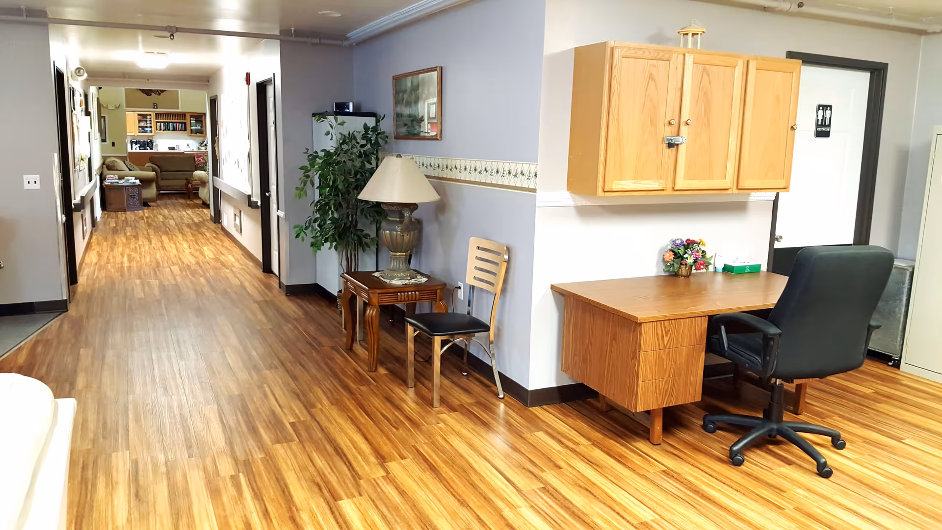 Interior hallway of a senior living facility with wood flooring, a desk with a black office chair, a wooden chair, a table with a lamp, a potted plant, and a seating area with couches visible at the end of the hallway. A restroom door is visible on the right side.