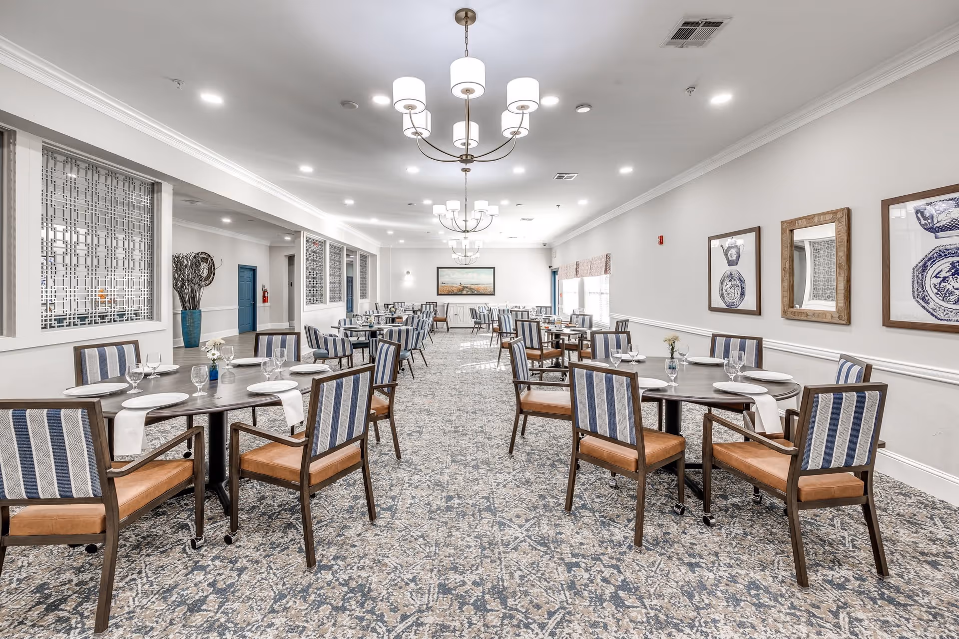 A spacious dining room with multiple round tables set with white plates, glasses, and napkins. Each table is surrounded by chairs with striped upholstery and wooden frames. The room features patterned carpet flooring, light gray walls adorned with framed artwork and a mirror, and modern chandeliers hanging from the ceiling. Large windows on the right side allow natural light to fill the space.