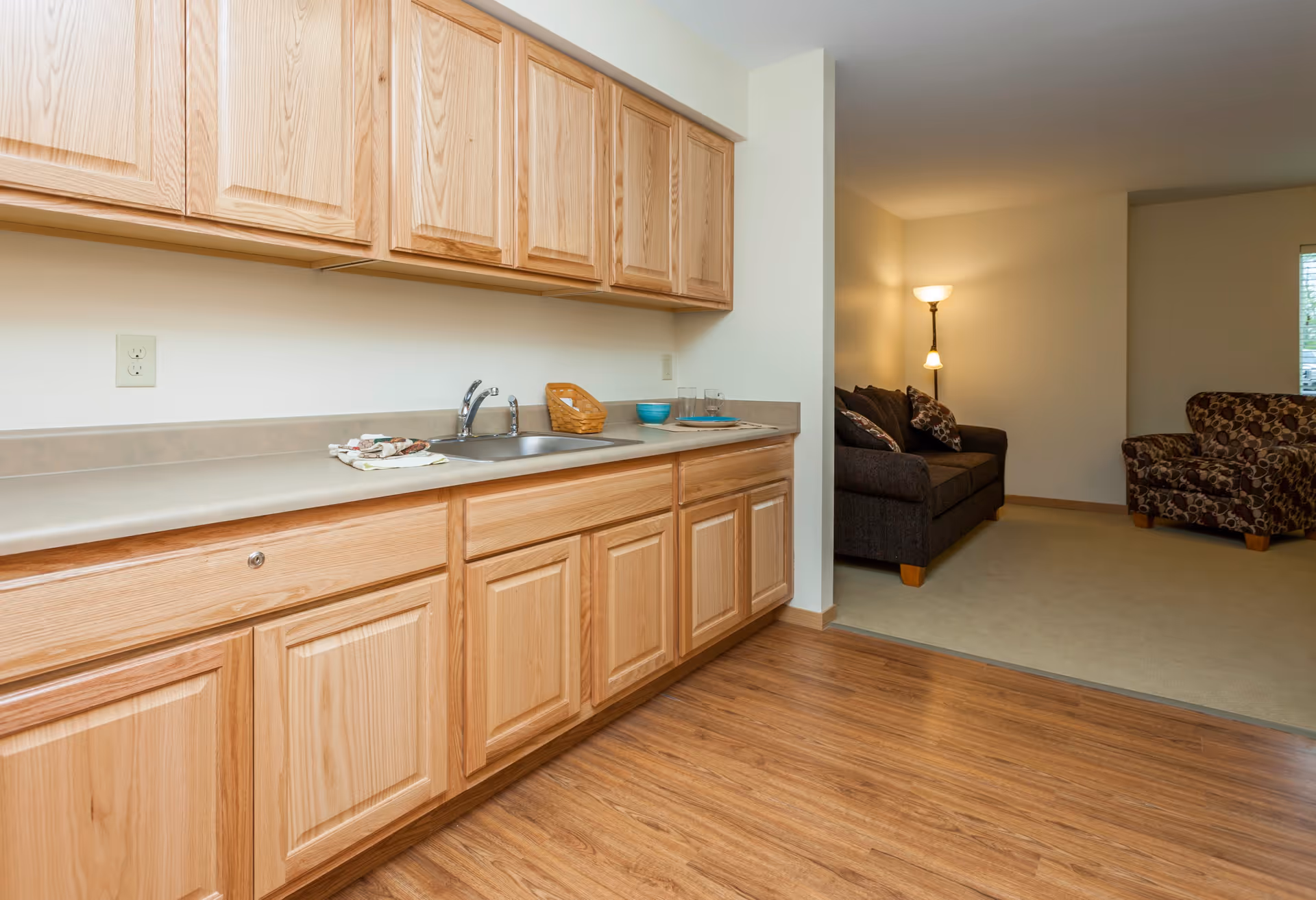 Interior view of a senior living facility showing a kitchenette with light wood cabinets, a countertop with a sink, and some dishes. Adjacent to the kitchenette is a living room area with a dark sofa, patterned armchair, a floor lamp, and a window with blinds.