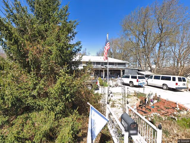 Exterior view of a senior living facility named Butterfly Haven with a large evergreen tree on the left, an American flag on a flagpole in front of the building, several white vans parked in the driveway, and a garden area with a white fence and mailbox in the foreground under a clear blue sky.
