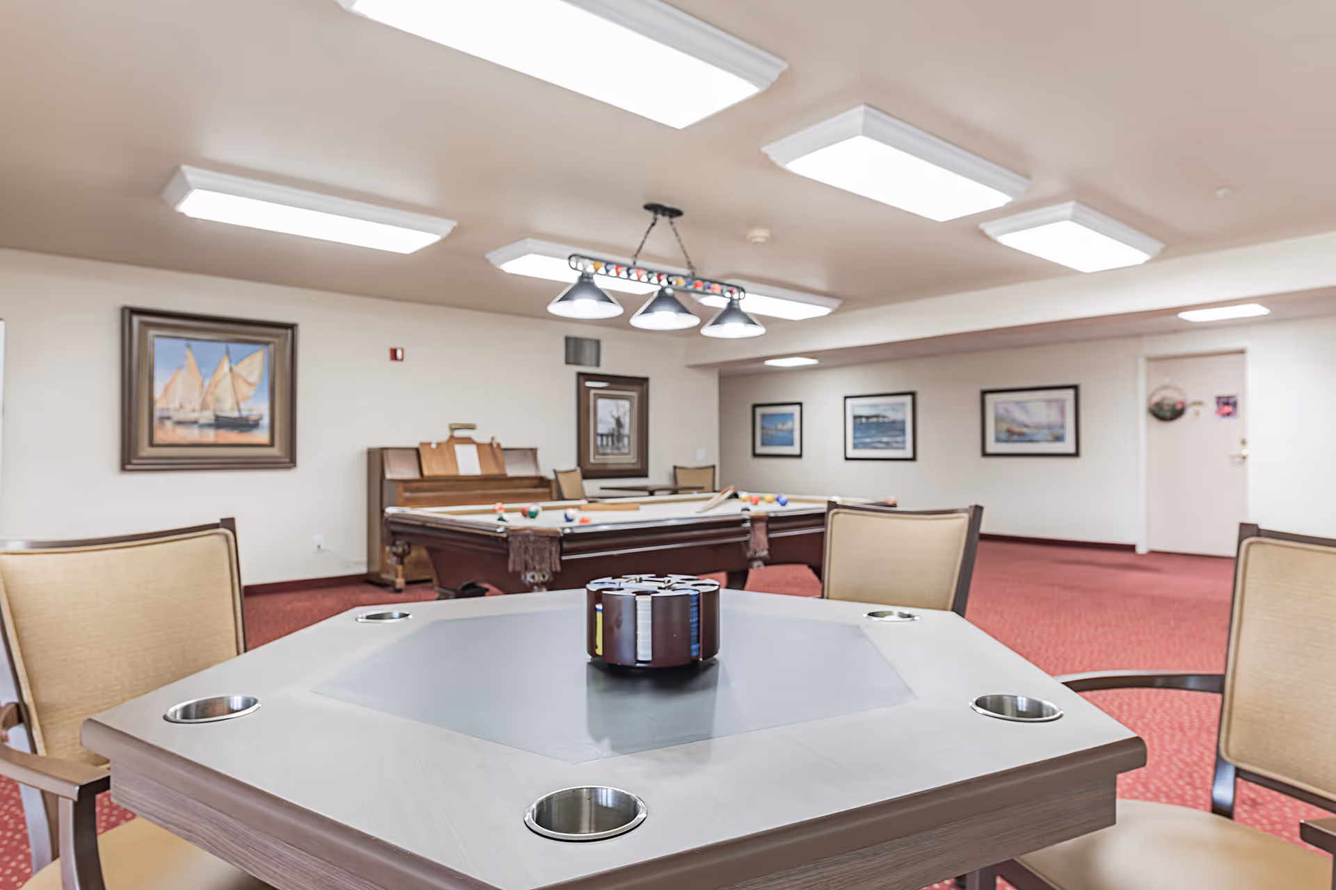 A recreational room with a card table in the foreground featuring cup holders and a poker chip holder. In the background, there is a pool table with billiard balls and cues, a piano, and framed paintings on the walls. The room has red carpet flooring and bright ceiling lights.