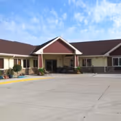 Exterior front view of a single-story senior living facility building with a covered entrance, beige walls, red gable, and a large concrete driveway in front under a partly cloudy sky.