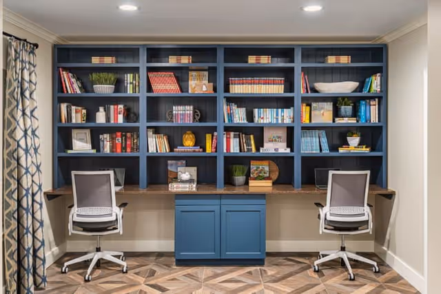 A study or work area with a built-in blue bookshelf filled with books and decorative items. Below the bookshelf is a long wooden desk with two modern office chairs on wheels positioned in front of it. The floor has a patterned wood design, and there is a curtain with a geometric pattern on the left side of the image.