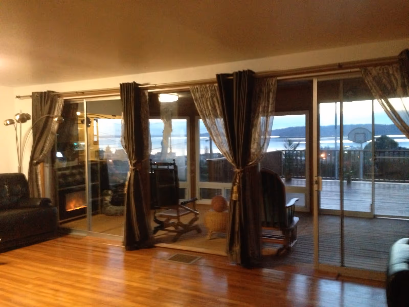 Interior view of a living room with wooden flooring, a black leather sofa on the left, a modern floor lamp, and large sliding glass doors with curtains leading to a balcony or patio area with rocking chairs and a view of water and hills in the background.