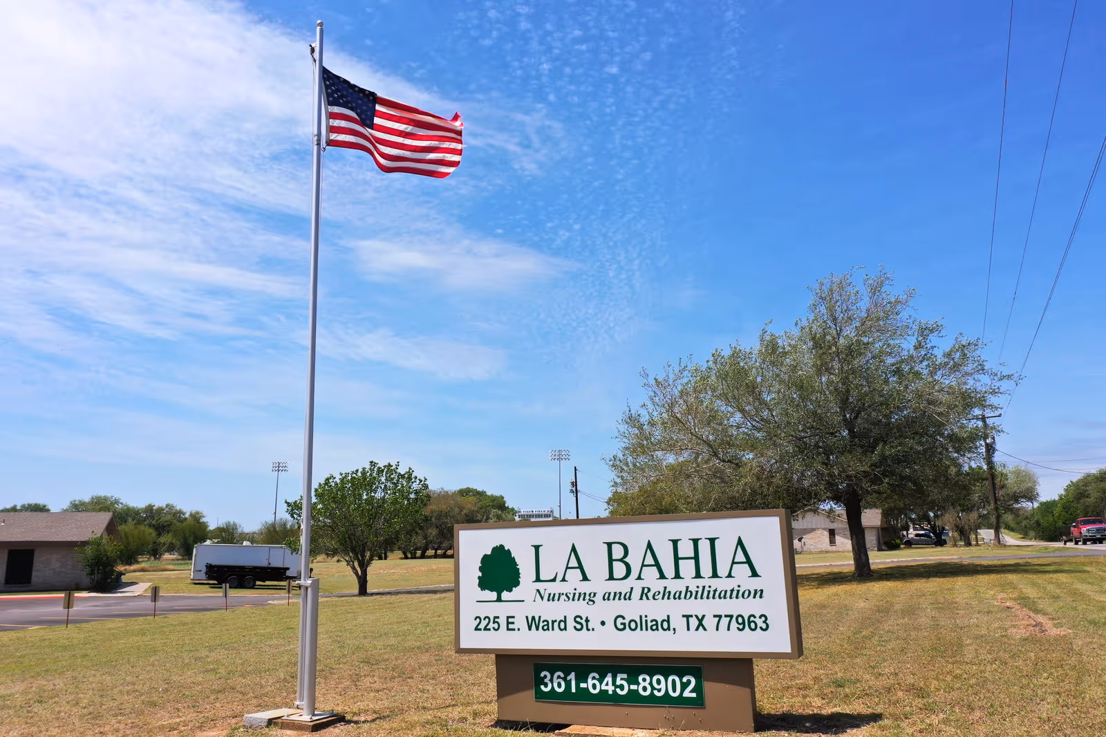 Outdoor view of La Bahia Nursing and Rehabilitation facility sign with an American flag on a flagpole nearby, trees, a truck, and a clear blue sky in the background.