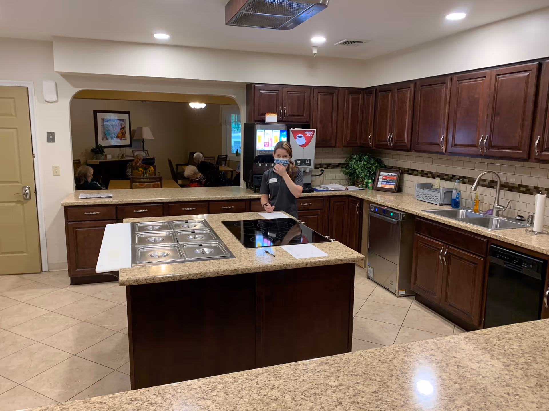 A kitchen area with dark wooden cabinets and beige countertops. A woman wearing a mask stands behind a counter with a stovetop and food warming trays. In the background, through an open archway, several elderly people are seated at tables in a dining or common area. The kitchen has a sink, dishwasher, and a beverage dispenser.
