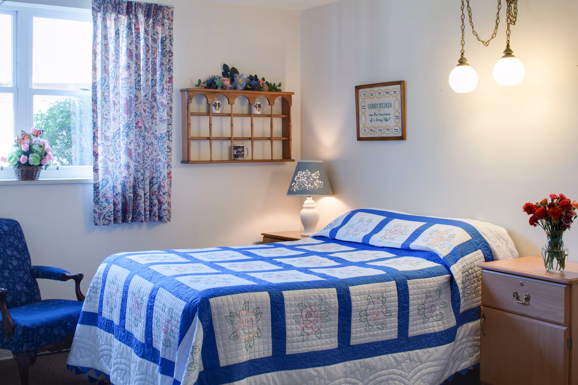 A cozy bedroom with a bed covered in a blue and white quilt featuring floral patterns. There is a wooden nightstand with a lamp and a vase of red flowers on the right side of the bed. A blue upholstered chair is positioned near a window with floral curtains. A wooden wall shelf with decorative items and a framed sign are mounted on the wall above the bed. Two hanging pendant lights provide warm lighting.