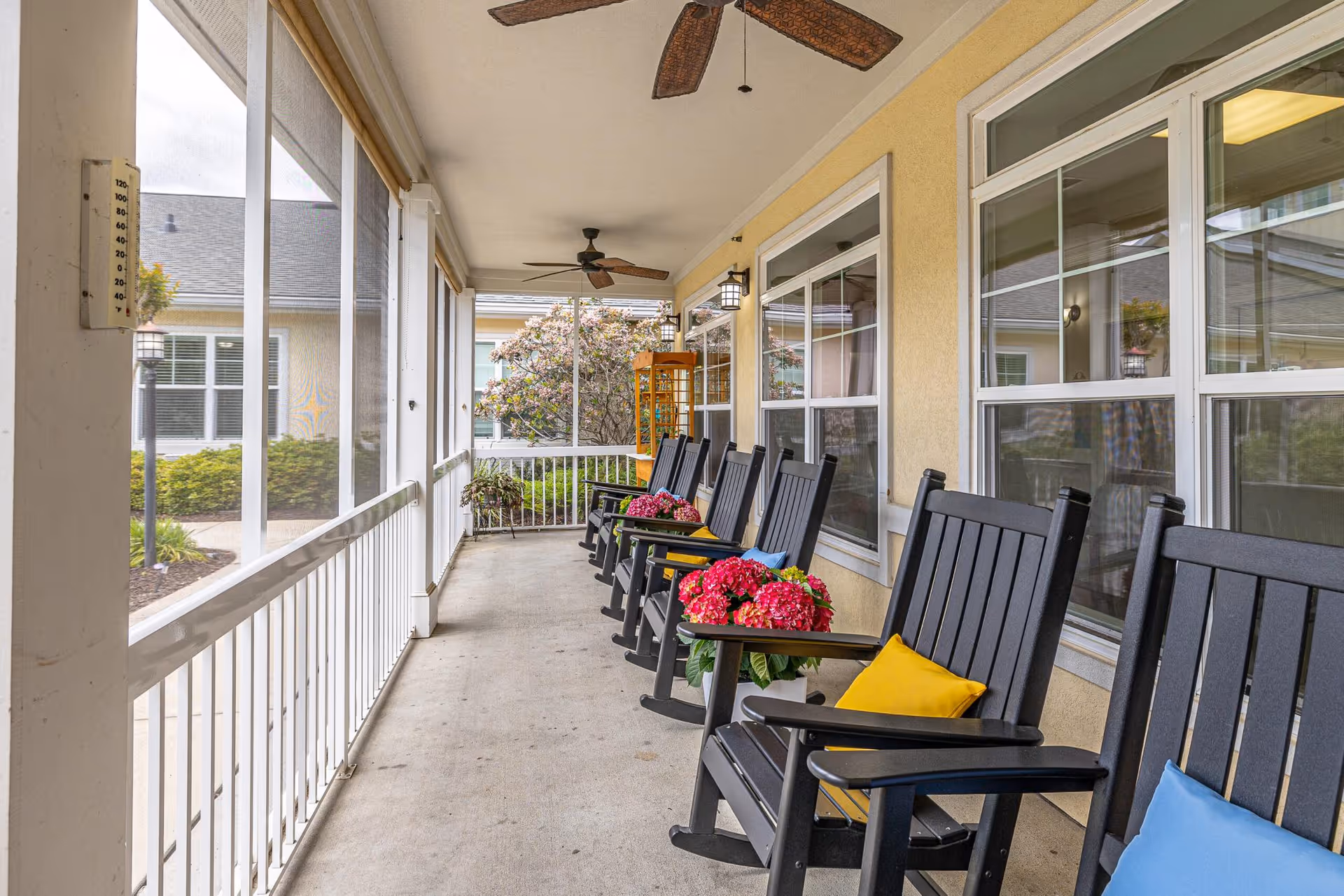 Screened porch with a row of black rocking chairs with colorful pillows and potted flowers, ceiling fans, and windows along the building wall.